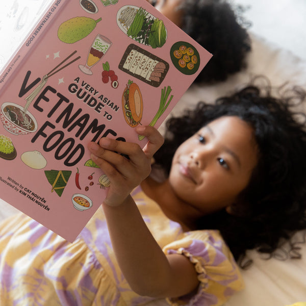 Child reading a kids book titled 'A Very Asian Guide to Vietnamese Food' on a bed with a friend.