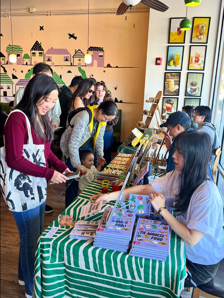 People interacting with a vendor at a table with books and stationery items in a store.